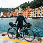 Das Bild zeigt Tobias Renggli mit seinem bepackten Gravel-bike auf einer Mole vor einem kleinen Ort am Meer.