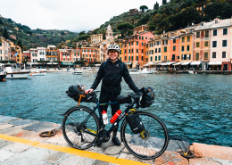 Das Bild zeigt Tobias Renggli mit seinem bepackten Gravel-bike auf einer Mole vor einem kleinen Ort am Meer.